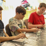 Grupa dzieciaków siedzi przy stole i uczestniczy w warsztatach plastycznych. Przed dziećmi leżą szklane elementy, które zamierzają pomalować.